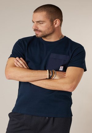 Homme aux cheveux courts et barbe portant un t-shirt bleu marine avec une poche, bras croisés, et plusieurs bracelets de perles, regardant vers la gauche.