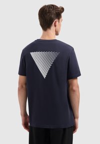 Man die met zijn rug naar de camera staat, gekleed in een marineblauwe t-shirt met een groot wit gestreept driehoekig ontwerp op de bovenrug.