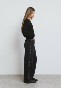 Un pull noir surdimensionné associé à un pantalon noir taille haute avec des boutons-pression en métal le long de la couture extérieure. Chaussures plates noires visibles.