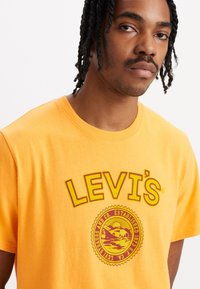 Sárga pamut póló, nagy kör alakú Levi's logóval bordó színben, a "LEVI'S" felirattal a kör felett és az "ESTABLISHED 1873" felirattal a kör belsejében.
