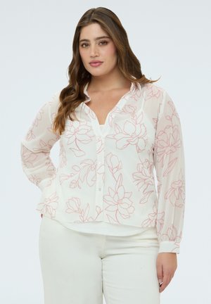 Frau mit langen braunen Haaren trägt eine weiße Bluse mit Knöpfen und rosa Blumenmuster sowie eine weiße Hose und steht vor einem hellen Hintergrund.