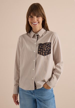 Femme souriante portant une chemise beige et blanche rayée à boutons avec une poche poitrine à imprimé léopard et un jean bleu, main dans la poche.
