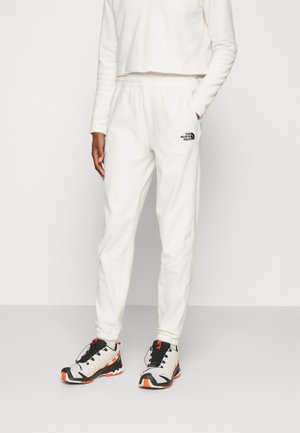 Osoba ubrana w białe spodnie dresowe z polaru z logo The North Face oraz białe, czarno-pomarańczowe buty sportowe, stojąca z jedną ręką w kieszeni.