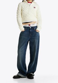 Personne portant un pull court en maille torsadée blanc, un jean large bleu avec des revers aux ourlets, des mocassins noirs, et une taille de pantalon Tommy Jeans visible.