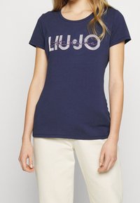 Kvinna iklädd en marinblå t-shirt med kort ärm och vit text samt beige byxor, stående mot en enfärgad ljus bakgrund.