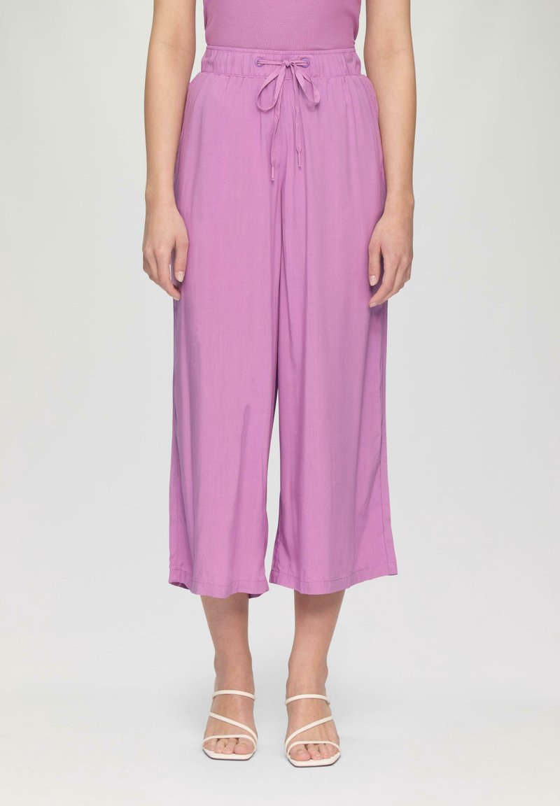 Pantalon large violet coupé au-dessus de la cheville avec taille à cordon, porté avec des sandales à talons blanches à brides sur fond uni.