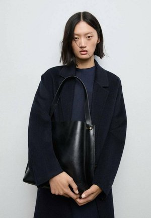 Borsa tote in pelle nera con un design minimalista, caratterizzata da un manico singolo e un sottile accento dorato vicino alla parte superiore. Texture liscia.