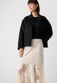 Black, wool-blend cropped jacket with a collared neckline and a single button closure, paired with a cream lace maxi skirt featuring floral patterns.