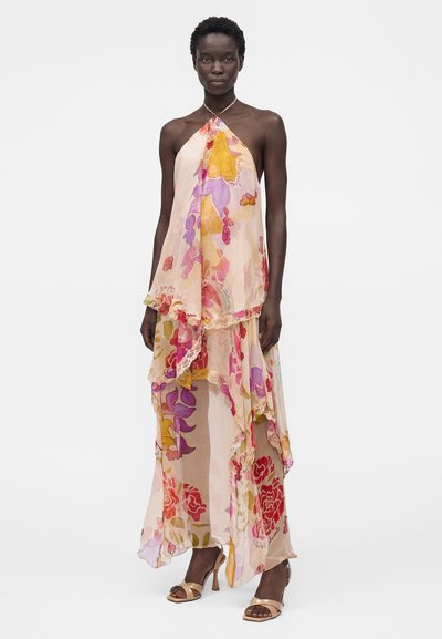 Γυναίκα που φοράει αμάνικο, διάφανο φλοράλ φόρεμα με στρωμένο ασύμμετρο τελείωμα, συνδυασμένο με γυναικείες ψηλές πέδιλα σε γυμνό χρώμα, στέκεται μπροστά σε λευκό φόντο.