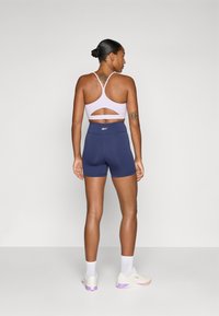 Sutien sport violet deschis cu detaliu tăiat și bretele subțiri; pantaloni scurți înalți bleumarin; adidași albi cu accente violet.