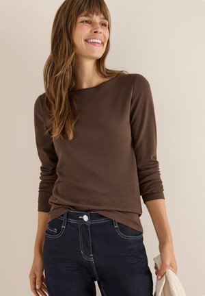Brauner, langärmeliger Top mit rundem Ausschnitt, kombiniert mit dunkelblauen Jeans mit Nahtdetails. Hält einen hellen Pullover.