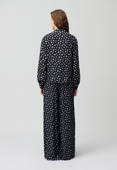 Completo in tessuto nero con motivo floreale bianco; include una blusa a maniche lunghe e pantaloni a gamba larga, entrambi con vestibilità rilassata e texture liscia.