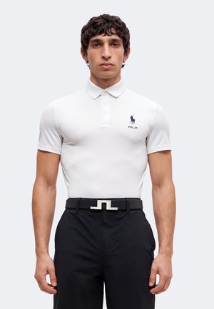 Homme aux cheveux courts et foncés portant un polo blanc ajusté avec un logo bleu et un pantalon noir avec une ceinture noire à boucle argentée.
