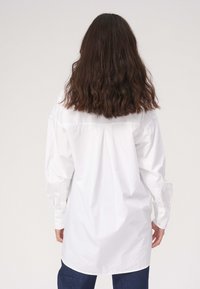 Hvid skjorte med lange ærmer, der har en bagrynke og rundet kant. Stoffet ser glat og bomuldsagtigt ud.