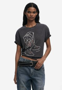 T-shirt graphique noir avec un design de botte de cowboy et des broderies colorées. Manches retroussées et coupe décontractée ; porté avec un jean bleu ample.
