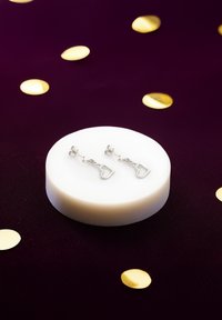 Orecchini d'argento a forma di cuore con una catena di collegamento, esposti su un piedistallo circolare bianco contro uno sfondo viola scuro con punti dorati.