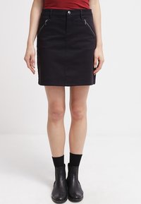 Schwarzer Jeansrock mit tailliertem Design, vorderen Reißverschlusstaschen und Gürtelschlaufen. Gekonnt kombiniert mit einem roten Oberteil und schwarzen Knöchelstiefeln.