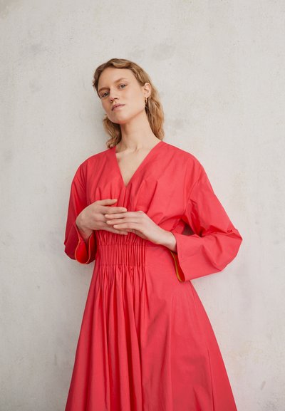 Vestido coral com decote em V, cintura franzida e mangas longas. Feito de um tecido leve, apresenta pregas subtis e uma textura suave.