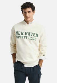 Crème kleurige sweatshirt met de tekst "NEW HAVEN SPORTS CLUB" in groene letters. Ribgebreide mouwen en zoom, relaxed fit, gecombineerd met donkere broeken.