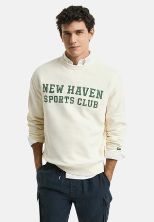 Sweatshirt de couleur crème avec "NEW HAVEN SPORTS CLUB" en texte vert. Manches et taille côtelées, coupe décontractée, associé à un pantalon sombre.
