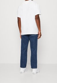 Camiseta de algodón blanca de manga corta, combinada con jeans de mezclilla azules. Los zapatos son zapatillas blancas. La vista muestra la parte posterior del conjunto.