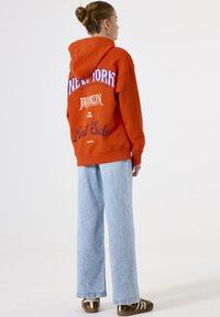 Woman with hair in bun wearing orange New York hoodie, light blue wide-leg jeans, and leopard print shoes with white straps, facing away.