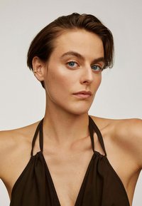 Top halter marrón con tela arrugada, con tirantes finos. La modelo tiene el cabello corto y ojos azul claro, y lleva un pendiente dorado.