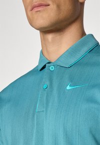 Turkizna polo majica z dvema gumboma, ovratnikom in Nike logotipom. Teksturirana tkanina s kontrastnim teal šivanjem ob ovratniku.