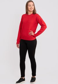 Roter gestrickter Pullover mit Rundhalsausschnitt und langen Ärmeln, kombiniert mit schwarzen Slim-Fit-Hosen und schwarzen Sneakers. Glatte Textur.