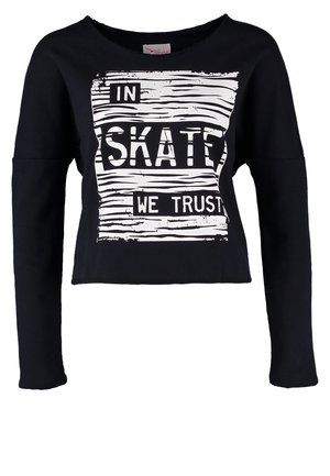 Person trägt einen schwarzen Sweatshirt mit dem Aufdruck „In Skate We Trust“, verblasste dunkle Jeans, marineblaue Sneakers und trägt einen schwarzen Rucksack auf einer Schulter.