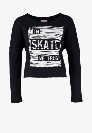 Osoba nosząca czarną bluzę z napisem „In Skate We Trust”, wyblakłe ciemne dżinsy, granatowe trampki i niosąca czarny plecak na jednym ramieniu.