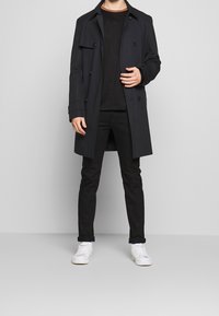 Black trench coat with double-breasted design, front pockets, and sleek fabric. Paired with a black shirt, black pants, and white sneakers.