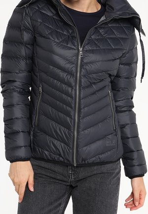 Veste matelassée noire avec coutures diagonales, fermeture éclair à l'avant et poches, portée sur une chemise noire et assortie à un jean foncé.