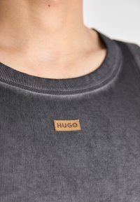 Pelēks ribotais tekstila tops ar apaļu kakla izgriezumu, kuram priekšā piestiprināta teksturēta brūna birka ar uzrakstu "HUGO".