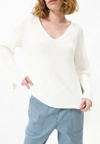 Witte, gestructureerde gebreide trui met een V-hals, lange mouwen met zijsplitten. Gecombineerd met blauw-witte gestreepte broek.