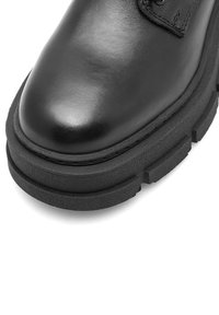 Pantof de piele neagră cu vârful rotunjit, suprafață superioară netedă și o talpă groasă din cauciuc texturat cu dispunere proeminentă a crampoanelor.