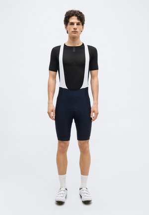 Jeune homme portant un maillot de cyclisme noir à manches courtes, un cuissard à bretelles bleu marine avec des bretelles blanches, des chaussettes blanches et des chaussures de cyclisme blanches, debout face à nous.