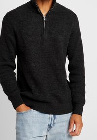 Dunkelgrauer, gerippter Strickpullover mit hohem Reißverschlusskragen. Der Stoff wirkt weich mit einer strukturierten Oberfläche und enganliegenden Bündchen sowie einem Bund.