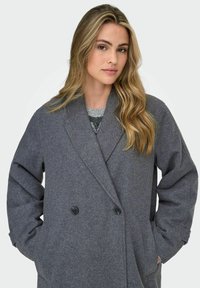 Young woman with long blonde hair wearing a gray double-breasted wool coat and a patterned sweater, hands in coat pockets, neutral background.