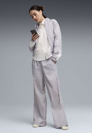 Mladá žena v svetlosivej súprave Puma a bielom tričku počúva slúchadlá, zatiaľ čo sa pozerá na svoj smartfón.