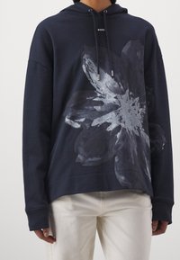 Mörkblå hoodies i mjukt material, med ett stort grått blommönster fram och en dragsko i huvan. Avslappnad passform med ribbade mudd.