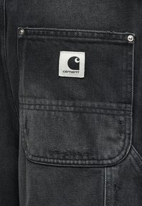 Τσέπη από μαύρο τζιν παντελόνι με λευκή υφασμάτινη ετικέτα με το λογότυπο Carhartt ραμμένη στο πάνω κέντρο της τσέπης.