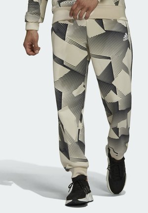 Homme portant un jogger beige avec des motifs géométriques noirs et des baskets Adidas noires, marchant vers l'avant.