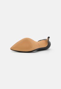 Chaussure plate noire à bout pointu avec une semelle flexible et une semelle extérieure en caoutchouc beige, présentant un design minimaliste sans matériel visible.