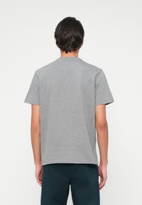 Vyras matomas iš nugaros, apsirengęs paprastu pilku trumpomis rankovėmis marškinėliais ir tamsiomis kelnėmis prieš baltą foną.