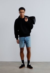 Άνδρας που φοράει μαύρο φούτερ, ανοιχτό μπλε σκισμένο τζιν σορτς, μαύρες κάλτσες και αθλητικά παπούτσια, κρατώντας μαύρη τσάντα στον ώμο μπροστά σε απλό φόντο.