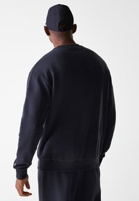 Sudadera navy, mezcla de algodón, cuello redondo, mangas largas, puños y dobladillo acanalados, textura suave, usada con una gorra oscura a juego.