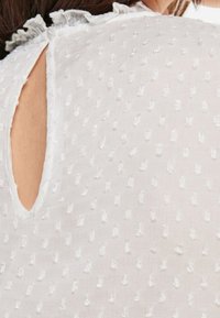 Top de tejido blanco con puntos texturizados, pequeña abertura en la parte trasera y detalle de cuello con volantes. Material suave y ligero con un diseño delicado.