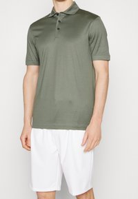 Olivgrön polo-skjorta med korta ärmar, treknappslåst, och slät struktur, bärs med vita shorts.