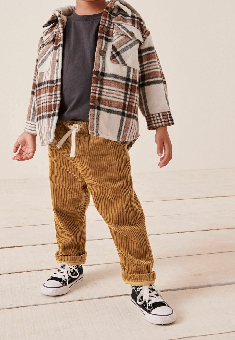 Enfant portant un pantalon en velours côtelé marron, des baskets noires, un t-shirt gris et une veste à carreaux marron et crème à boutons, debout sur un sol en bois.